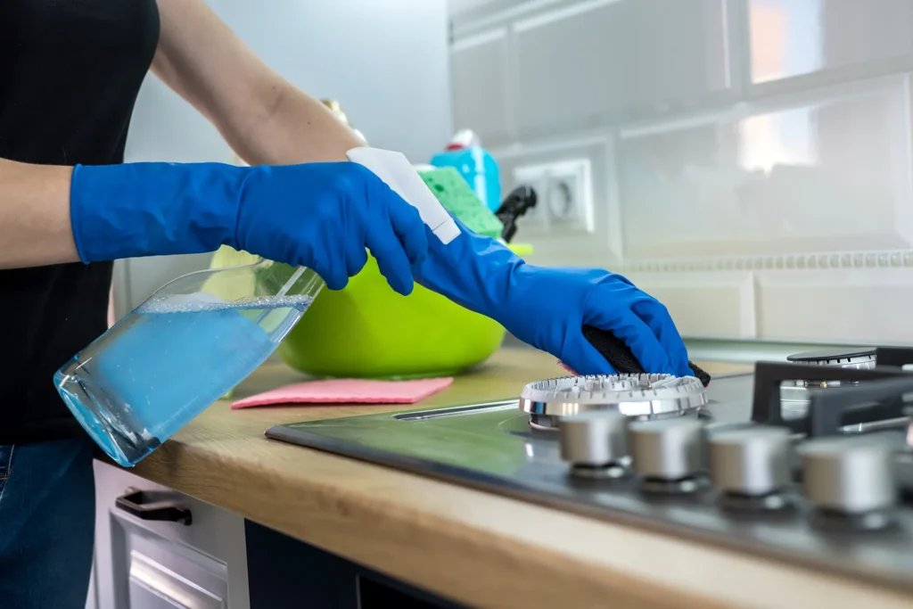 Women in blue gloves cleaning a steel stove counter top with a blue spray bottle
